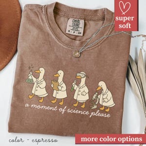May include: Espresso-colored t-shirt featuring a whimsical design of four ducks in lab coats, each engaged in a scientific activity. The phrase "a moment of science please" is printed below the ducks. The shirt has a crew neck and short sleeves.