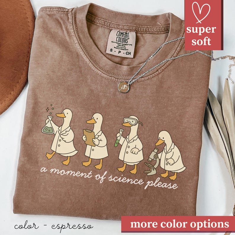 May include: Espresso-colored t-shirt featuring a whimsical design of four ducks in lab coats, each engaged in a scientific activity. The phrase "a moment of science please" is printed below the ducks. The shirt has a crew neck and short sleeves.