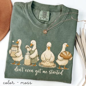 May include: Moss green Comfort Colors t-shirt with a humorous illustration of ducks baking, accompanied by the phrase "don't even get me started." A silver necklace with a pendant is placed on the shirt.