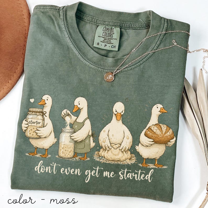 May include: Moss green Comfort Colors t-shirt with a humorous illustration of ducks baking, accompanied by the phrase "don't even get me started." A silver necklace with a pendant is placed on the shirt.