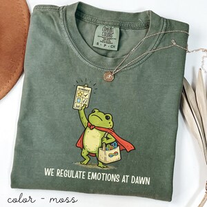May include: Moss green Comfort Colors t-shirt featuring a cartoon frog superhero holding a schedule and a shopping bag. The text "WE REGULATE EMOTIONS AT DAWN" is printed below the frog. A gold necklace with a pendant is draped on the shirt.