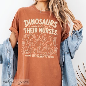 May include: A rust-colored t-shirt featuring a cream-colored graphic of various dinosaurs dressed as medical professionals. The text reads: DINOSAURS didn't listen to THEIR NURSES. and we all know WHAT HAPPENED TO THEM. The design includes medical icons.