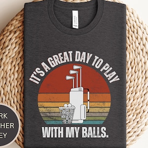 May include: Dark heather grey t-shirt with a vintage-style graphic of a golf bag and golf balls. The text reads "It's a great day to play with my balls."