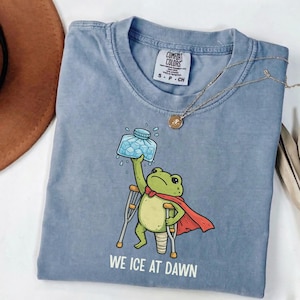 May include: A blue Comfort Colors t-shirt with a cartoon frog superhero holding an ice pack and using crutches. The text "WE ICE AT DAWN" is printed at the bottom. A gold necklace is draped across the shirt.