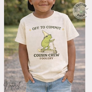 K&ouml;nnte beinhalten: Ein cremefarbenes T-Shirt mit einem Cartoon-Frosch auf einem Roller, der einen Zaubererhut tr&auml;gt. Der Text "OFF TO COMMIT" und "COUSIN CREW FOOLERY" sind &uuml;ber und unter der Frosch-Illustration aufgedruckt. Das T-Shirt wird von einem Kind getragen.