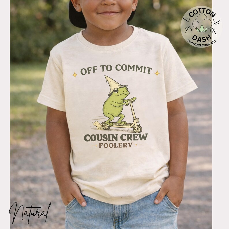 K&ouml;nnte beinhalten: Ein cremefarbenes T-Shirt mit einem Cartoon-Frosch auf einem Roller, der einen Zaubererhut tr&auml;gt. Der Text "OFF TO COMMIT" und "COUSIN CREW FOOLERY" sind &uuml;ber und unter der Frosch-Illustration aufgedruckt. Das T-Shirt wird von einem Kind getragen.