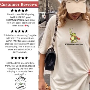 May include: Cream-colored t-shirt featuring a cartoon frog superhero holding a sign, with the text "WE REGULATE EMOTIONS AT DAWN." The shirt is part of a clothing line, likely a graphic tee.