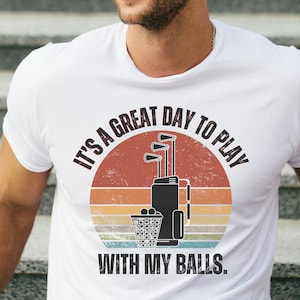 May include: White t-shirt with a vintage-style graphic of golf clubs and a golf bag. The text reads "It's a great day to play with my balls."