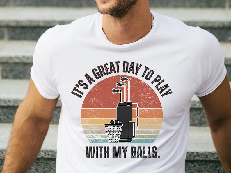 May include: White t-shirt with a vintage-style graphic of golf clubs and a golf bag. The text reads "It's a great day to play with my balls."