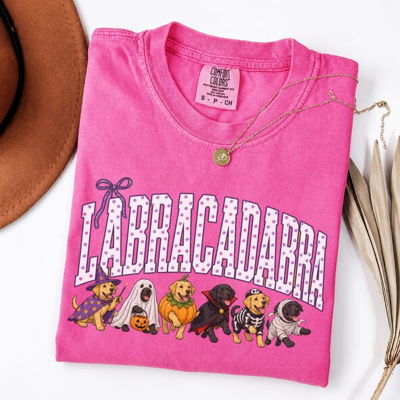 May include: A bright pink t-shirt featuring the text LABRACADABRA in white with purple polka dots. Below the text are seven dogs in various Halloween costumes, including a wizard, ghost, pumpkin, vampire, skeleton, and mummy, with a gold necklace.