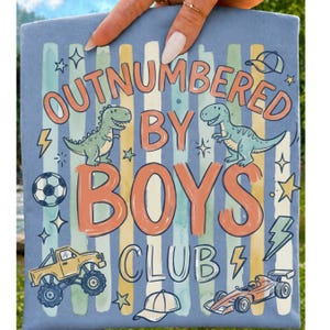 May include: A light blue graphic tee with the text "OUTNUMBERED BY BOYS CLUB". The design includes cartoon dinosaurs, a soccer ball, a monster truck, a race car, lightning bolts, stars, and baseball caps. The background features vertical stripes in various colours.