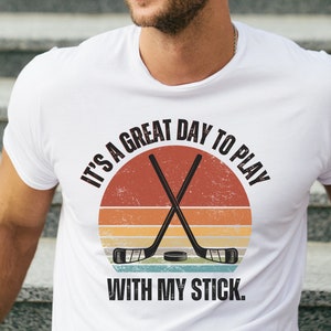 K&ouml;nnte beinhalten: Wei&szlig;es T-Shirt mit einer Grafik im Vintage-Stil, auf der "It's a great day to play with my stick." steht. Die Grafik zeigt einen Hockeyschl&auml;ger und einen Puck.
