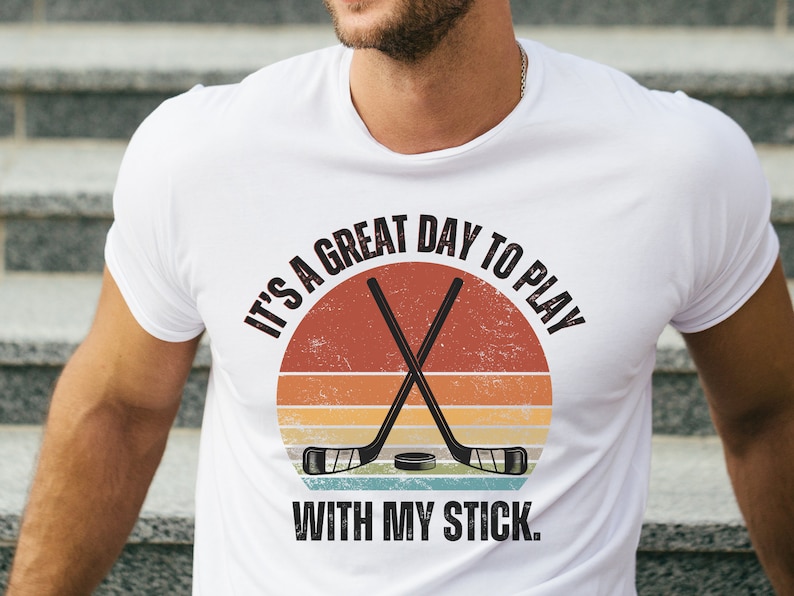 K&ouml;nnte beinhalten: Wei&szlig;es T-Shirt mit einer Grafik im Vintage-Stil, auf der "It's a great day to play with my stick." steht. Die Grafik zeigt einen Hockeyschl&auml;ger und einen Puck.