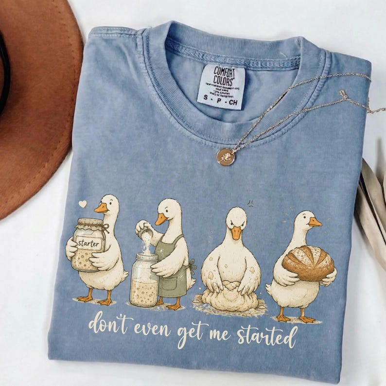 May include: A pale blue Comfort Colors t-shirt with a humorous illustration of ducks baking. The ducks are shown with jars, aprons, and a loaf of bread. The text "don't even get me started" is printed below the ducks. A silver necklace is also visible.