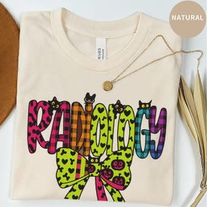 Puede incluir: Camiseta color crema con la palabra "RADIOLOGY" en letras coloridas y estampadas. El diseño incluye ilustraciones de gatos negros y un lazo verde con detalles de corazones y calabazas. También se ve un collar dorado con un colgante.