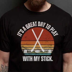 K&ouml;nnte beinhalten: Ein schwarzes T-Shirt mit einer Grafik im Vintage-Stil von gekreuzten Hockeyschl&auml;gern &uuml;ber einem Sonnenuntergang. Der Text lautet "Es ist ein gro&szlig;artiger Tag, um mit meinem Schl&auml;ger zu spielen."