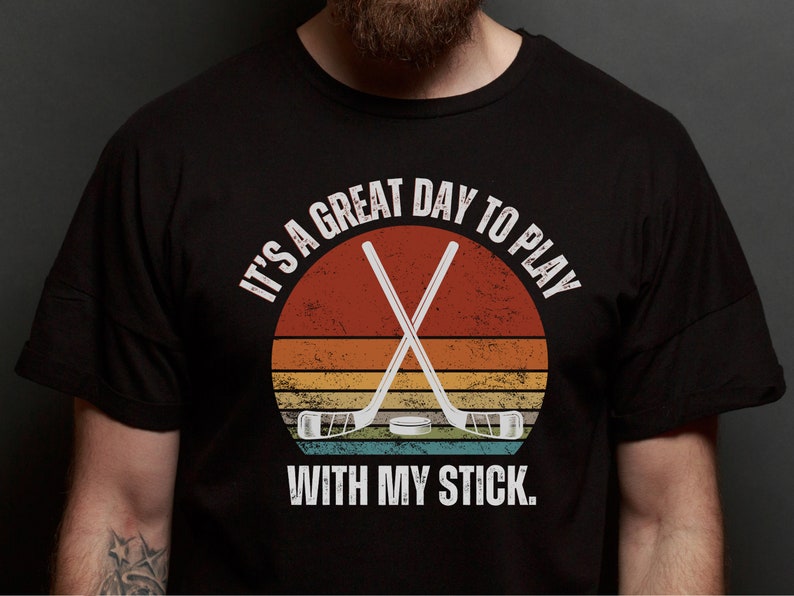 K&ouml;nnte beinhalten: Ein schwarzes T-Shirt mit einer Grafik im Vintage-Stil von gekreuzten Hockeyschl&auml;gern &uuml;ber einem Sonnenuntergang. Der Text lautet "Es ist ein gro&szlig;artiger Tag, um mit meinem Schl&auml;ger zu spielen."