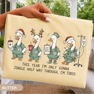 May include: A light yellow blanket featuring four cartoon ducks dressed as nurses in green scrubs. The text reads: THIS YEAR I'M ONLY GONNA JINGLE HALF WAY THROUGH, I'M TIRED. The ducks wear holiday accessories like Santa hats, reindeer antlers, and Christmas lights.