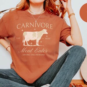 May include: A rust-colored t-shirt with a white cow graphic and the text "CARNIVORE EAT BEEF Meat Eater SAVING THE HUMANS".