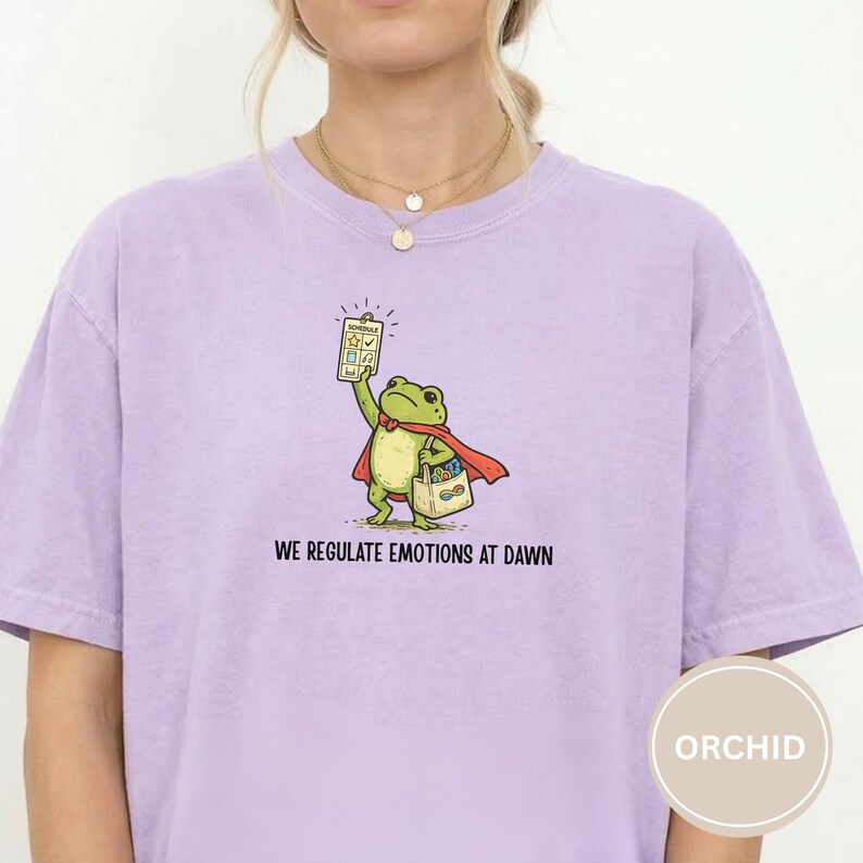 May include: A light purple t-shirt featuring a cartoon frog wearing a red cape and holding a checklist. The frog carries a bag with colorful items. The text "WE REGULATE EMOTIONS AT DAWN" is printed below the frog.