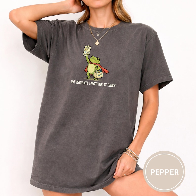 May include: Dark grey t-shirt featuring a cartoon frog superhero with a red cape and a sign. The frog is holding a box and a sign that reads "WE REGULATE EMOTIONS AT DAWN". The shirt is a relaxed fit.