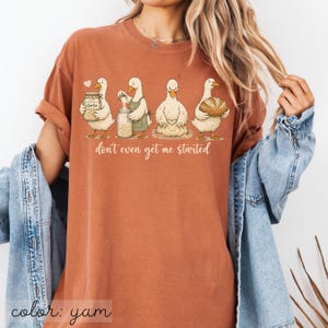 May include: A yam-coloured t-shirt with a whimsical illustration of four ducks involved in baking. The ducks are depicted with jars, aprons, and a loaf of bread, with the phrase "don't even get me started" below.