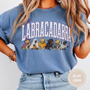 May include: A blue crewneck t-shirt featuring the text LABRACADABRA in purple polka-dot letters above a graphic of six dogs in various Halloween costumes, including a wizard, ghost, pumpkin, vampire, skeleton, and mummy, all in a cartoon style.