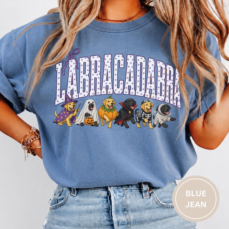 May include: A blue crewneck t-shirt featuring the text LABRACADABRA in purple polka-dot letters above a graphic of six dogs in various Halloween costumes, including a wizard, ghost, pumpkin, vampire, skeleton, and mummy, all in a cartoon style.