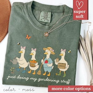 Puede incluir: Camiseta verde musgo con una ilustración caprichosa de cuatro patos jardineros, cada uno con una herramienta u objeto diferente. El texto "just doing my gardening stuff" está debajo de los patos. Un collar dorado está sobre la camiseta.