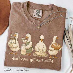 May include: Espresso-coloured t-shirt with a cartoon illustration of four geese baking. The geese are shown with jars, dough, and a loaf of bread. The text "don't even get me started" is printed below the geese. A silver necklace is also visible.