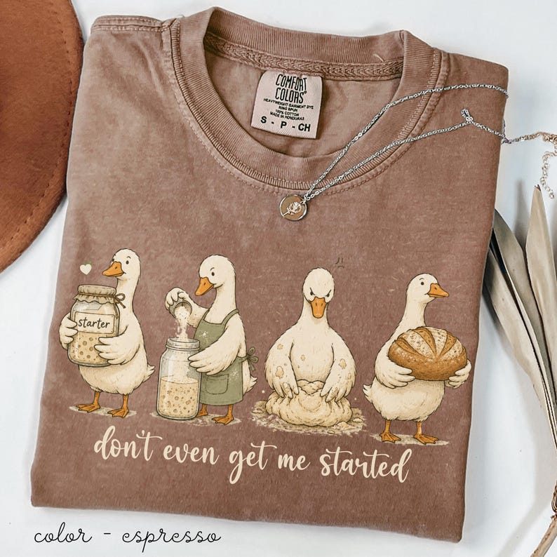 May include: Espresso-coloured t-shirt with a cartoon illustration of four geese baking. The geese are shown with jars, dough, and a loaf of bread. The text "don't even get me started" is printed below the geese. A silver necklace is also visible.