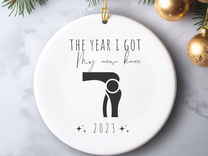 Op de afbeelding: Witte kerstbal van keramiek met zwarte tekst die "Het jaar dat ik mijn nieuwe knie kreeg, 2023" luidt en een zwart silhouet van een knie.