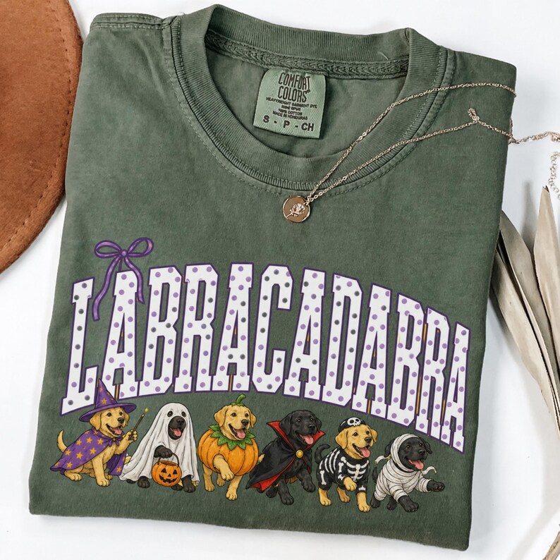 May include: An olive green crew neck t-shirt featuring the text "LABRACADABRA" in white with purple polka dots. Below the text are six Labrador dogs dressed in Halloween costumes: a wizard, a ghost, a pumpkin, a vampire, a skeleton, and a mummy.