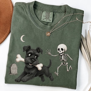May include: An olive green crew neck t-shirt featuring a graphic print of a black terrier dog holding a bone in its mouth, running away from a small human skeleton. A tombstone marked R.I.P. and a crescent moon are also printed on the fabric.