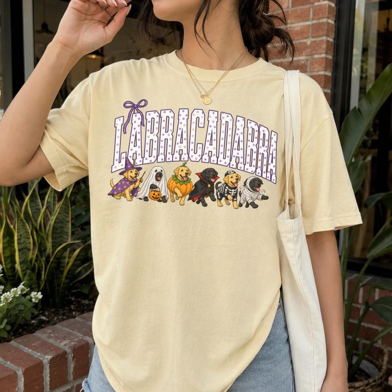 May include: A pale yellow t-shirt featuring the text LABRACADABRA in purple polka-dot letters. Below the text are six dogs in Halloween costumes: a wizard, a ghost, a pumpkin, a vampire, a skeleton, and a mummy, all in a row.