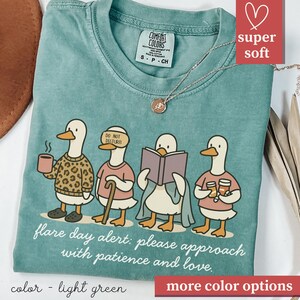 May include: Light green Comfort Colors t-shirt featuring a whimsical illustration of four ducks. Each duck is depicted with unique accessories, including a coffee cup, a blindfold, a book, and drinks. The text "flare day alert: please approach with patience and love" is printed below the ducks.
