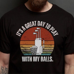 May include: A black t-shirt with a vintage-style graphic of a golf bag with clubs and golf balls. The text reads "It's a great day to play with my balls."
