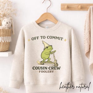 K&ouml;nnte beinhalten: Ein meliertes naturfarbenes Sweatshirt mit dem Text "OFF TO COMMIT" und "COUSIN CREW FOOLERY". Die Grafik zeigt einen gr&uuml;nen Frosch auf einem Roller mit einem Zaubererhut. Das Wort "heather natural" ist kursiv geschrieben.