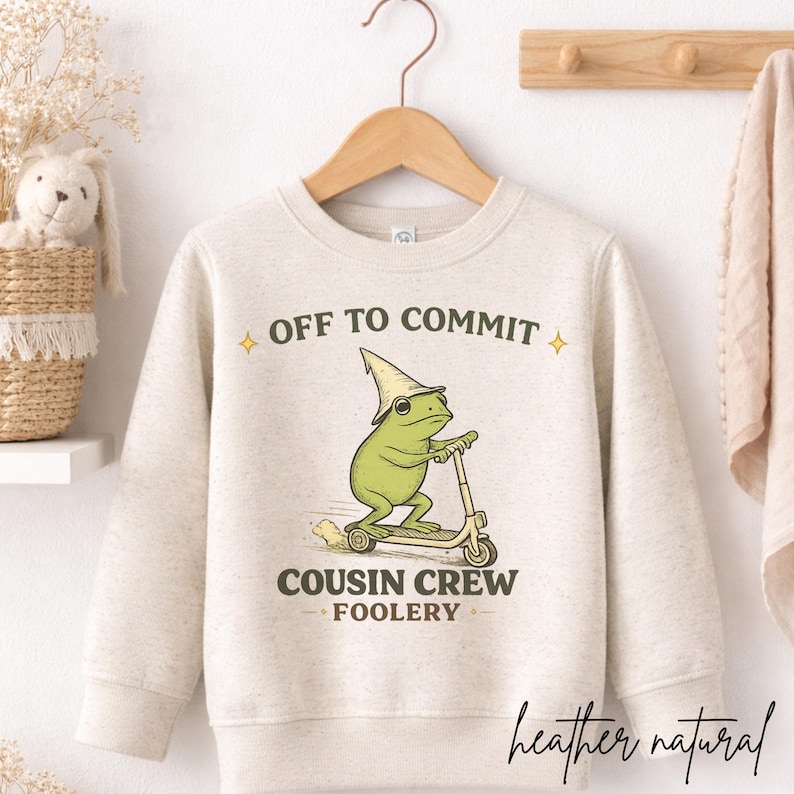 K&ouml;nnte beinhalten: Ein meliertes naturfarbenes Sweatshirt mit dem Text "OFF TO COMMIT" und "COUSIN CREW FOOLERY". Die Grafik zeigt einen gr&uuml;nen Frosch auf einem Roller mit einem Zaubererhut. Das Wort "heather natural" ist kursiv geschrieben.