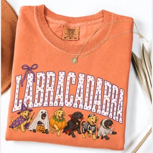 May include: An orange short-sleeved t-shirt featuring the text "LABRACADABRA" in white with purple polka dots. Below the text are six Labrador dogs in Halloween costumes: a wizard, a ghost, a pumpkin, a vampire, a skeleton, and a mummy.