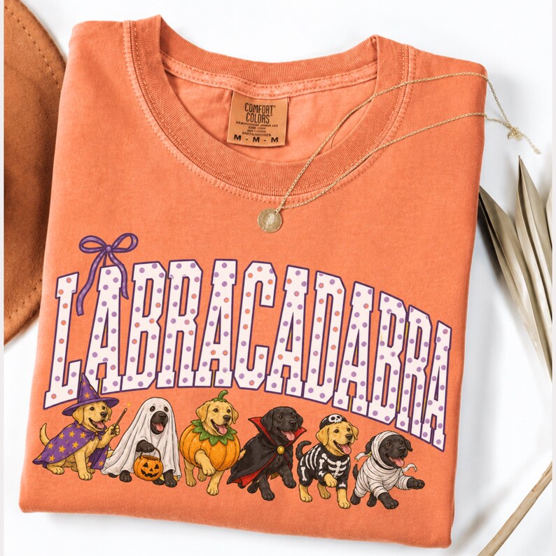 May include: An orange short-sleeved t-shirt featuring the text "LABRACADABRA" in white with purple polka dots. Below the text are six Labrador dogs in Halloween costumes: a wizard, a ghost, a pumpkin, a vampire, a skeleton, and a mummy.