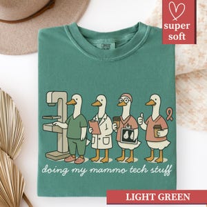 May include: Light green t-shirt with a crew neck and the words "doing my mammo tech stuff" in a script font. The graphic features four cartoon ducks dressed as medical professionals. The shirt is labeled "super soft" and "light green".