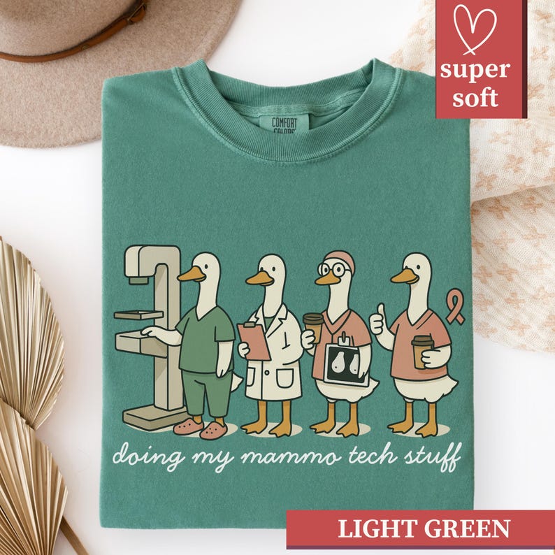 May include: Light green t-shirt with a crew neck and the words "doing my mammo tech stuff" in a script font. The graphic features four cartoon ducks dressed as medical professionals. The shirt is labeled "super soft" and "light green".