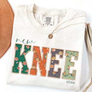 Pode incluir: Camiseta creme com as palavras "new KNEE" em um design floral. As letras são delineadas com pequenas contas douradas. O texto "EST.2026" está abaixo da palavra "KNEE". Um colar de ouro está sobre a camiseta.