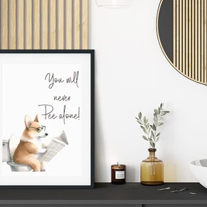 May include: Framed art print featuring a watercolor illustration of a corgi wearing glasses and reading a newspaper while sitting on a toilet. The text above the corgi reads, "You will never Pee alone!" The print is in a black frame.
