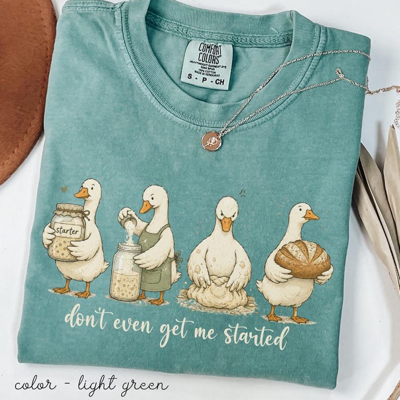 May include: Light green Comfort Colors t-shirt with a humorous illustration of geese holding jars and bread. The text "don't even get me started" is below the geese. A silver necklace with a pendant is also visible. The shirt is a small size.