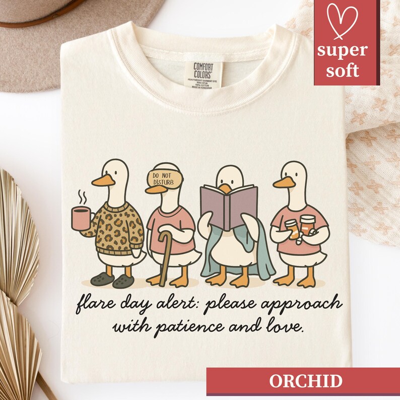 May include: Cream-colored t-shirt with a whimsical illustration of four ducks, each with unique attire and accessories. The ducks are depicted with a coffee cup, a blindfold, a book, and drinks. The text "flare day alert: please approach with patience and love" is printed below the ducks.