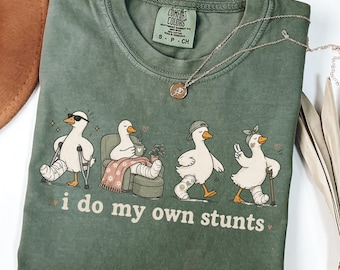 May include: A moss green short-sleeve t-shirt featuring a graphic of four white ducks with injuries, including casts and crutches. The text below the ducks reads, "i do my own stunts." The shirt has a Comfort Colors label at the inner neckline.