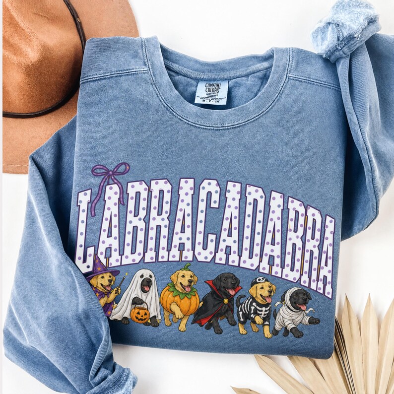 May include: A blue crewneck sweatshirt featuring the text LABRACADABRA in white with purple polka dots. Below the text are illustrations of six Labrador dogs in Halloween costumes, including a wizard, ghost, pumpkin, vampire, skeleton, and mummy.