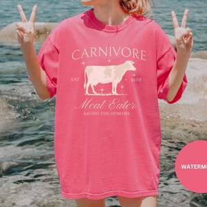 May include: Pink t-shirt with a white cow graphic and the text "CARNIVORE Eat Beef Meat Eater Saving the Humans" on the front. The shirt is being worn by a person standing in front of a body of water.